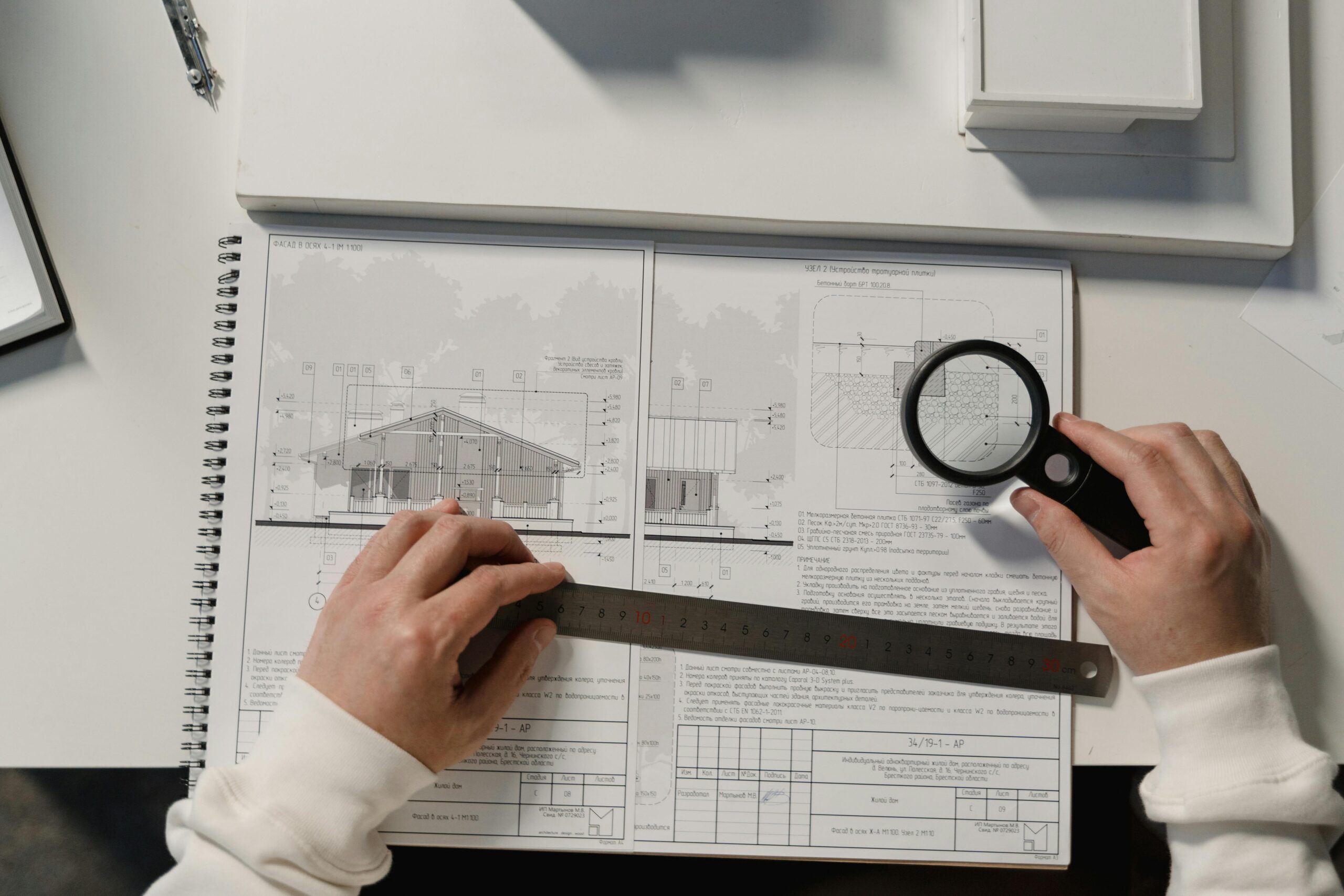 Hands holding a ruler and magnifying glass over architectural floor plans on a white table.